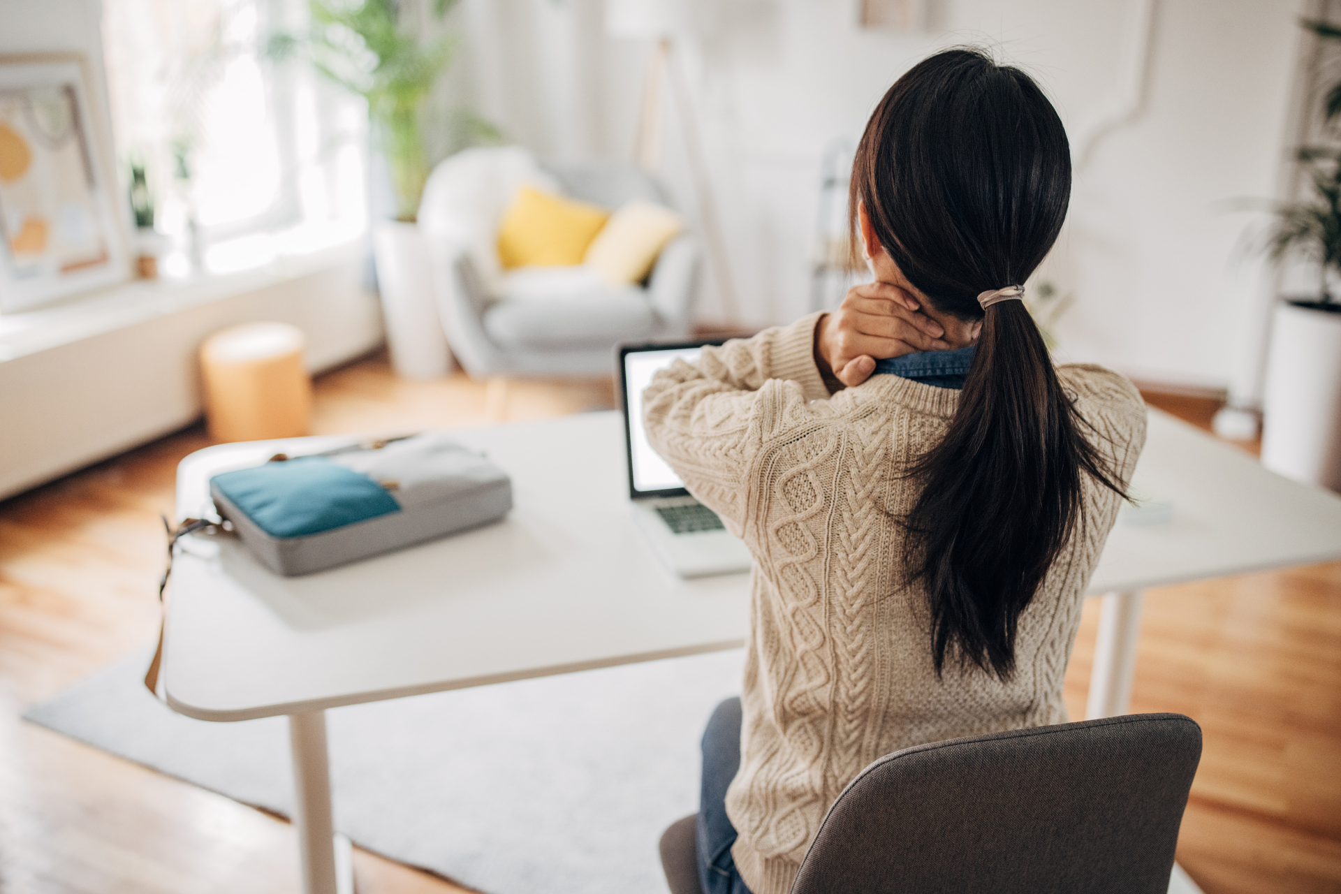 Woman sitting at a desk holding the back of her neck in pain while using a laptop