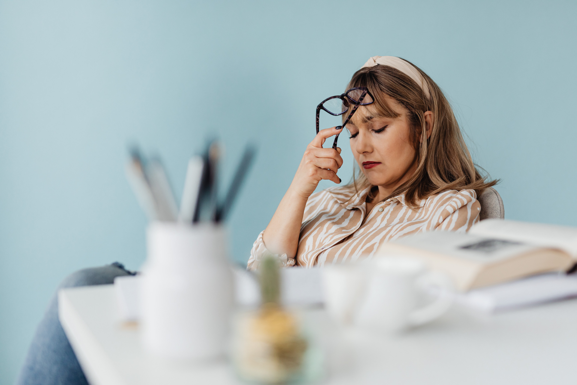 Woman sitting at a desk with eyes closed, pressing glasses to her forehead