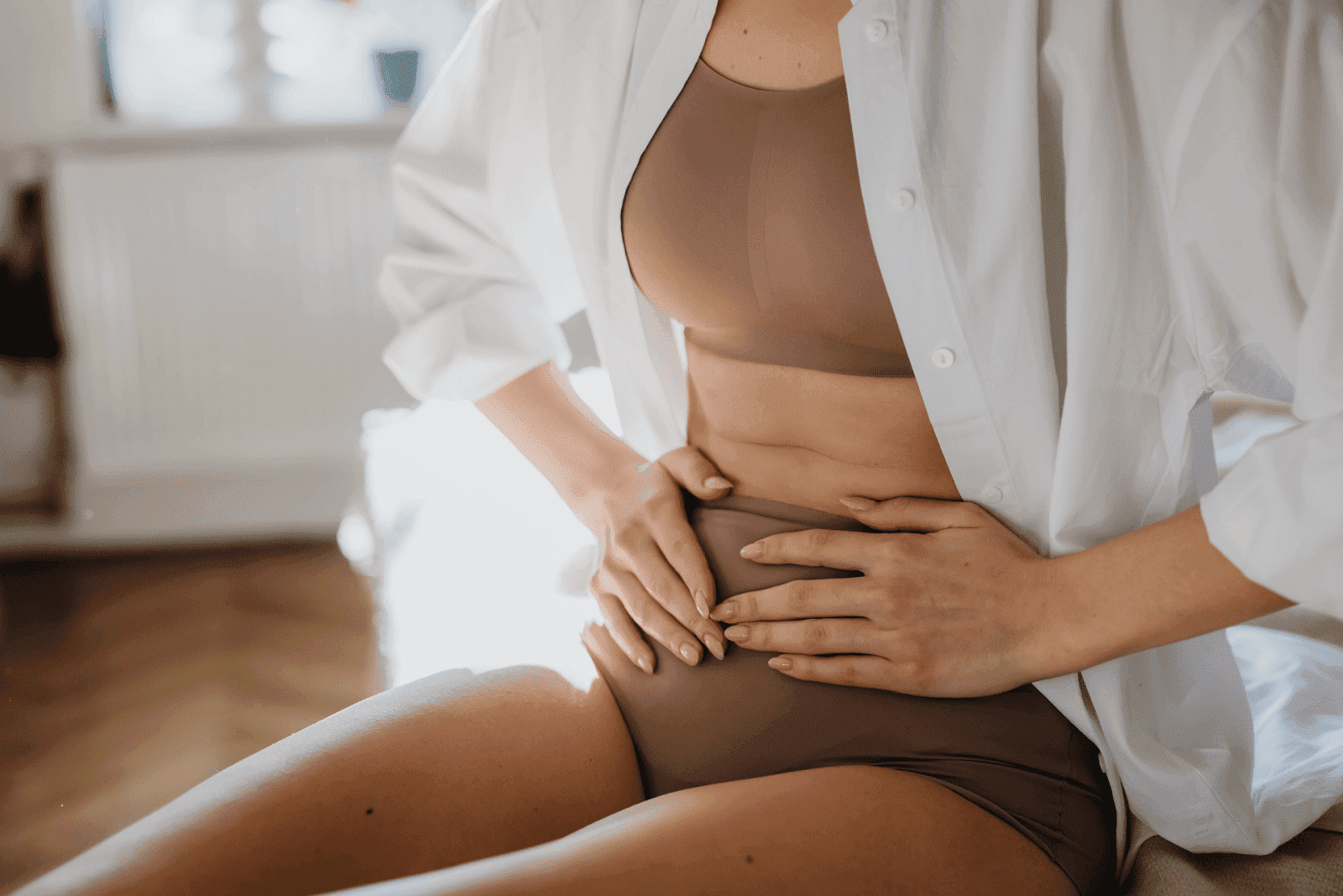 Woman sitting in underwear and shirt, pressing her stomach with both hands in pain.