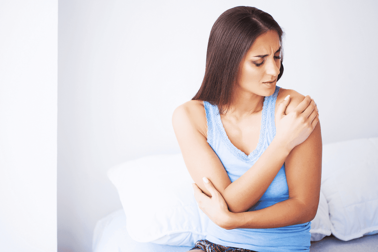 Woman sitting on a bed holding her arm and shoulder in pain, appearing uncomfortable.