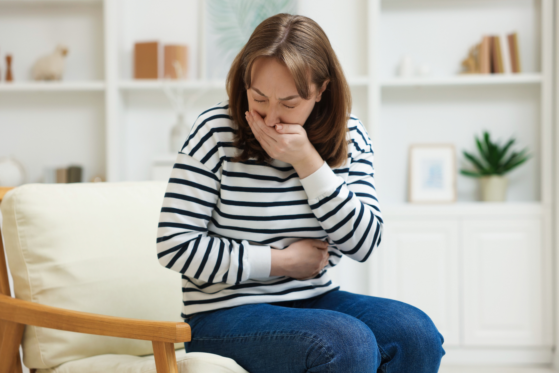 Woman sitting on a chair holding her stomach and covering her mouth, appearing nauseous
