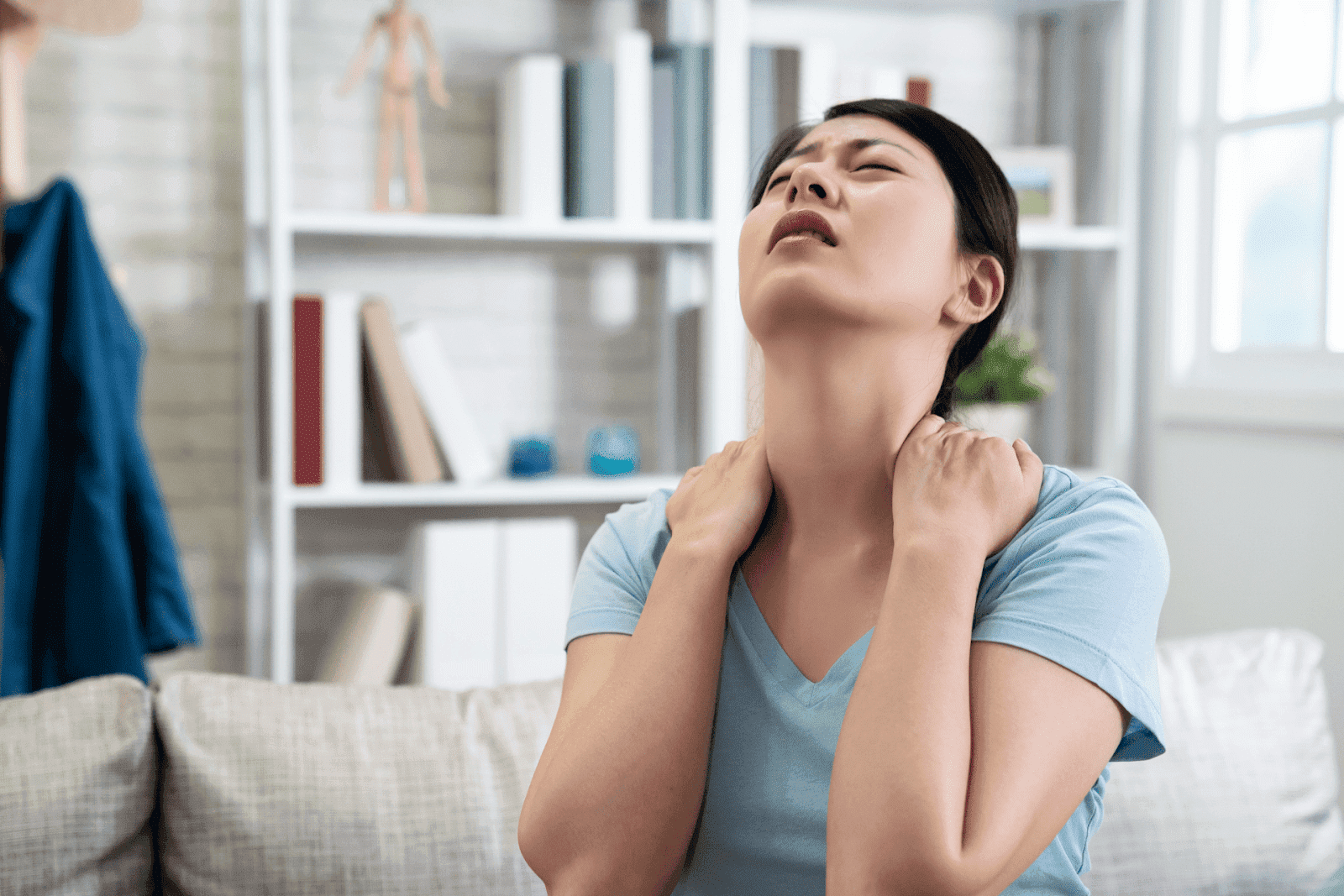 Woman sitting on a couch, pressing her neck and shoulders in discomfort.
