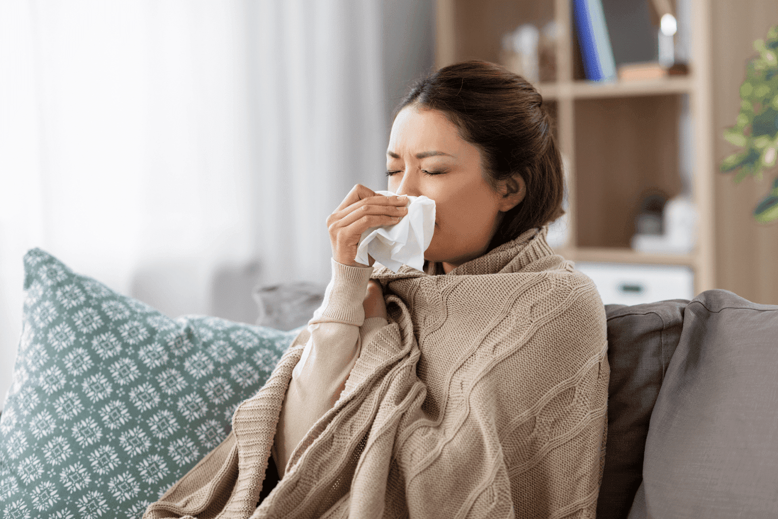 Woman sitting on a couch, wrapped in a blanket, sneezing into a tissue