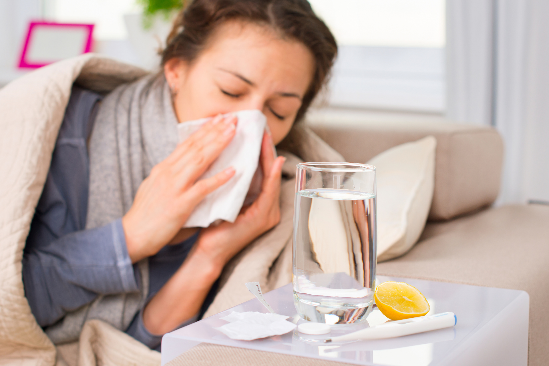Woman sitting on a couch wrapped in a blanket, blowing her nose with a tissue, with a glass of water and lemon on a table beside her, appearing sick at home