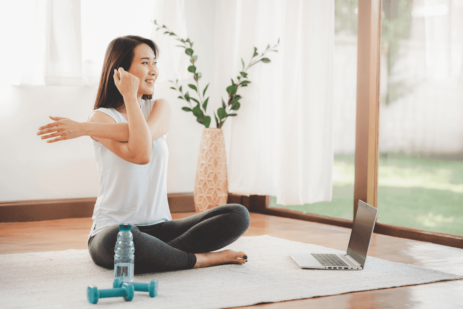 Woman sitting on a mat at home stretching her arm, with a water bottle, dumbbells, and a laptop nearby