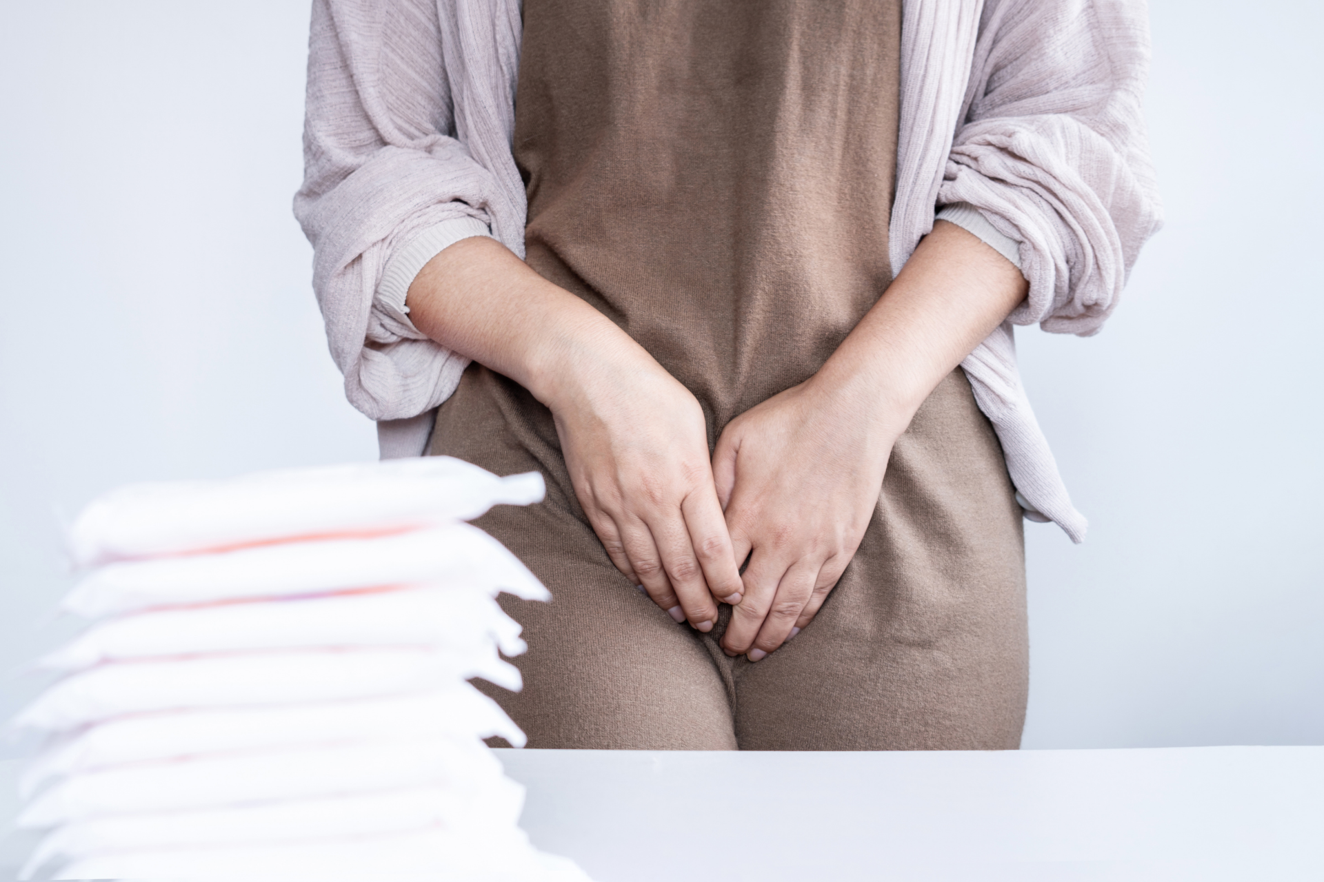 Woman sitting with hands clasped over her lower abdomen, with sanitary pads on a table nearby