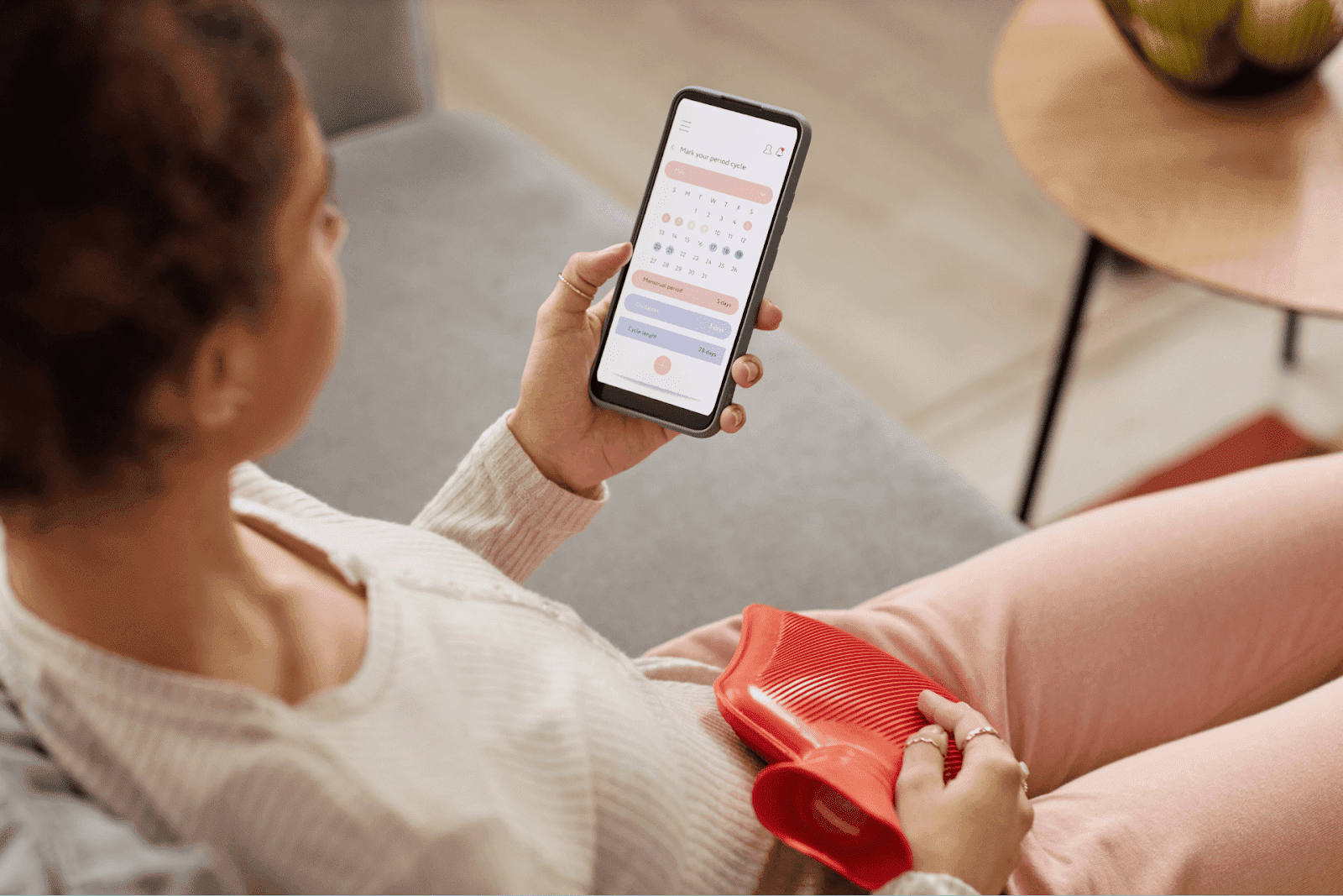 Woman tracking menstrual cycle on smartphone while holding a red hot water bottle on her stomach.