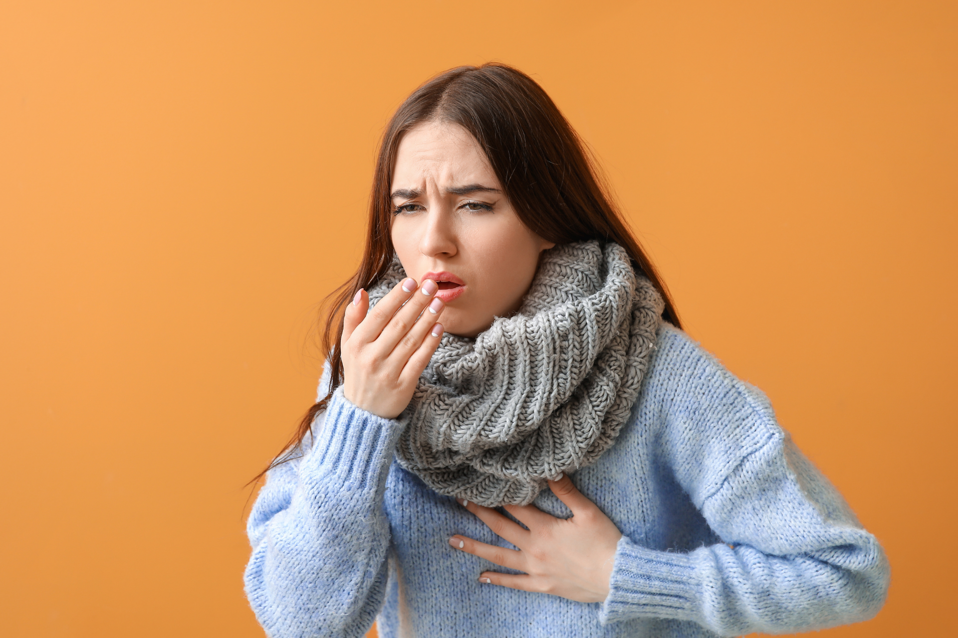 Woman wearing a scarf and sweater, coughing and holding her chest against an orange background