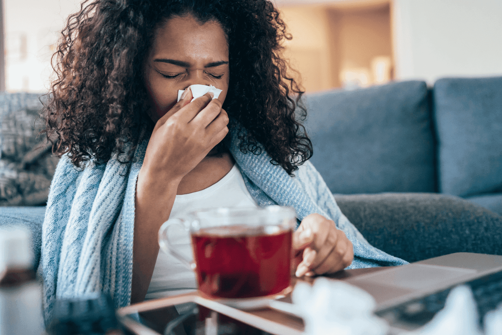 Woman wrapped in a blanket, holding a tissue and drinking hot tea, appearing sick.