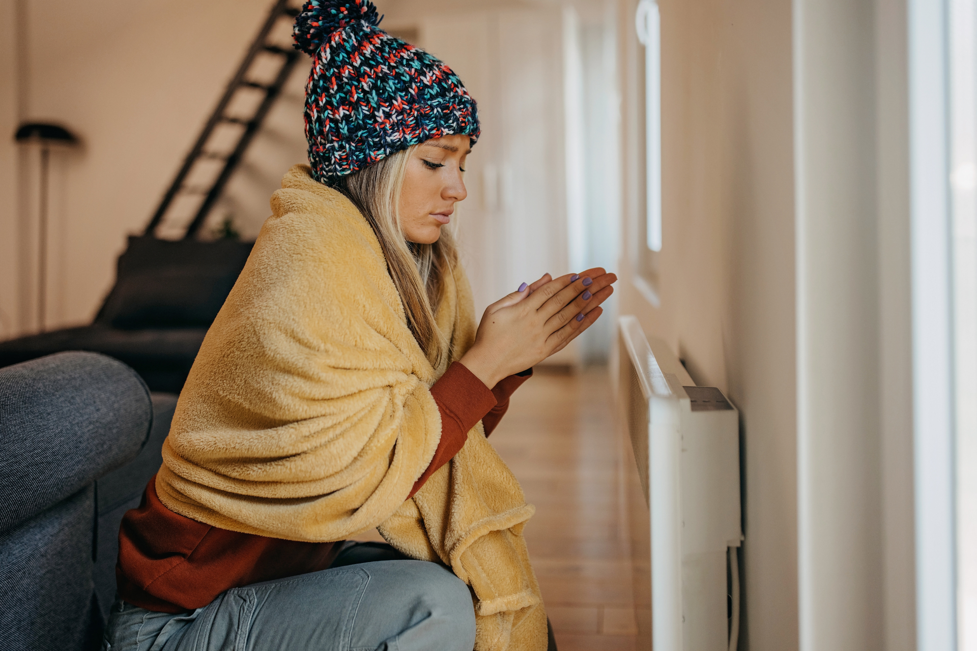 Woman wrapped in a blanket warming her hands near a heater indoors