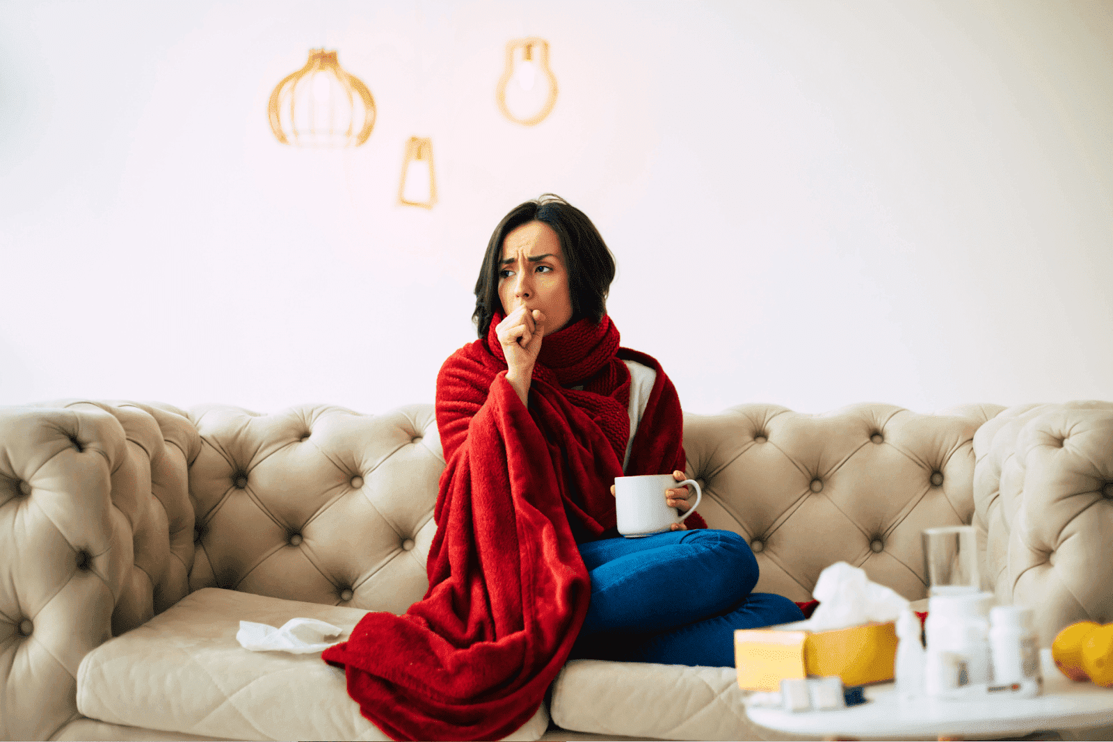 Woman wrapped in a red blanket, coughing while sitting on a couch with medicines on the table