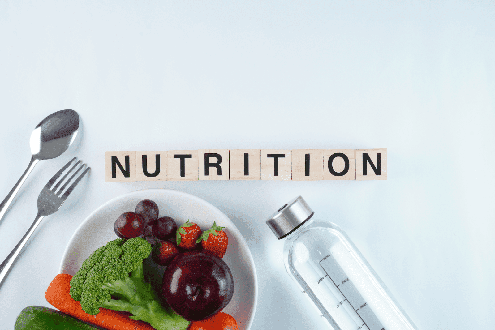 Word ‘Nutrition’ spelled with wooden blocks beside a plate of fruits, vegetables, and a water bottle.