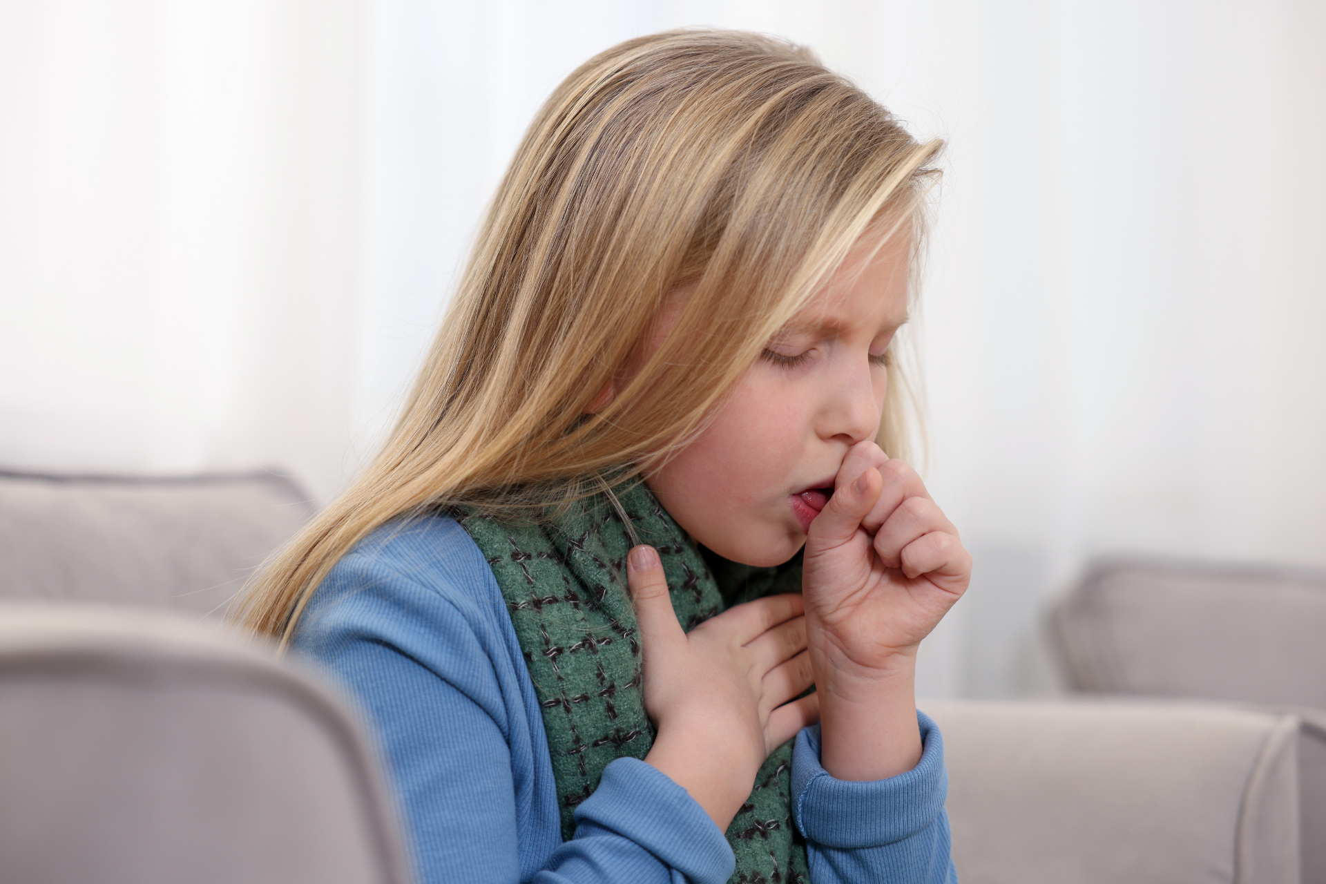 Young child coughing with a hand over their mouth and the other on their chest, appearing unwell while sitting indoors
