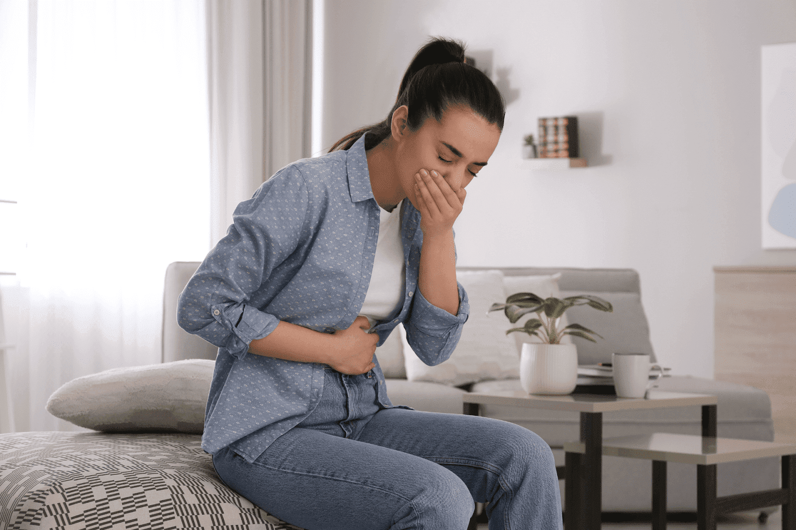 Young woman sitting on bed holding her stomach and covering her mouth, appearing nauseous.