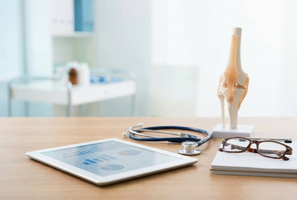 Medical desk with a knee joint model, stethoscope, tablet with charts, and glasses on a notebook.