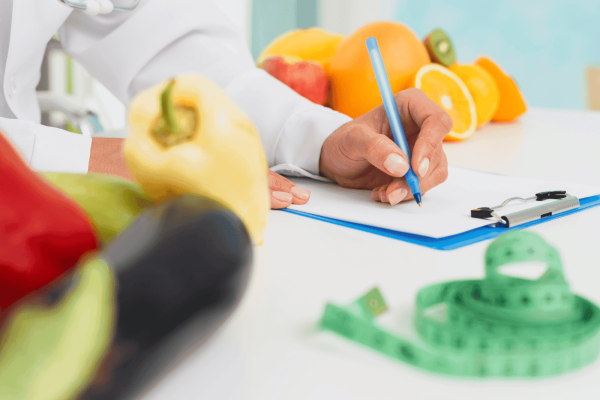 Nutritionist writing on a clipboard surrounded by fresh fruits and vegetables with a measuring tape on the table.