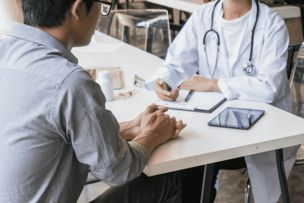 Patient consulting with a doctor in a white coat at a desk with a clipboard and tablet during a medical appointment.