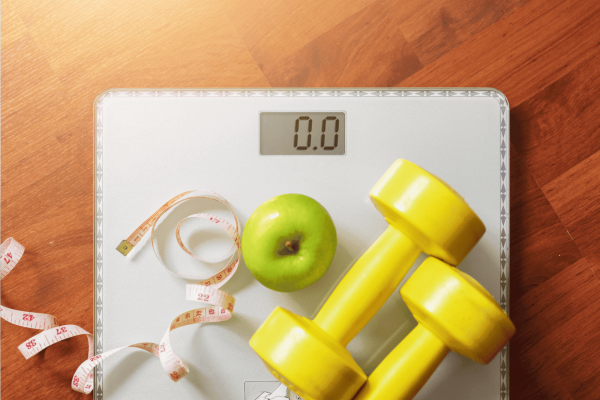 Scale with an apple, measuring tape, and yellow dumbbells symbolizing fitness and weight management.