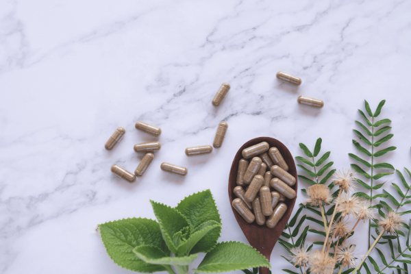 Herbal supplement capsules on a marble surface with green leaves and flowers.