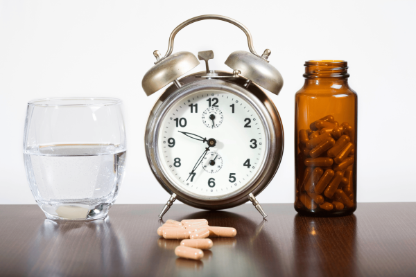 Clock, glass of water, and medicine bottle with capsules on a table.