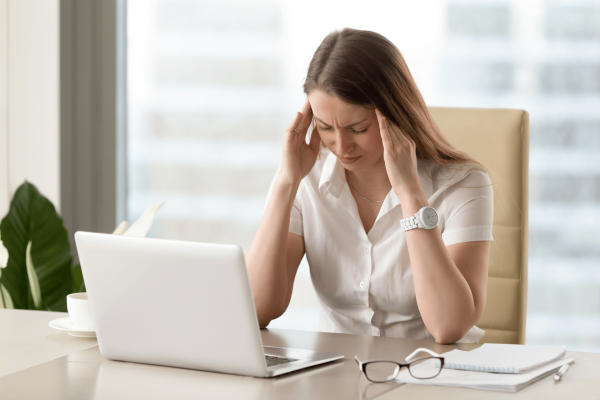 Woman sitting at a desk with a laptop, holding her head in pain or stress.