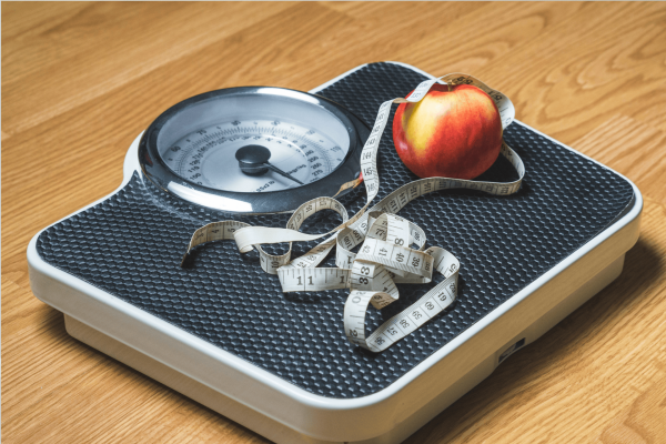 Bathroom scale with a measuring tape and an apple on top, symbolizing weight management.
