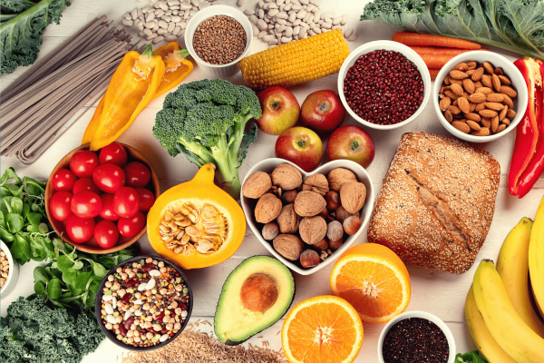 Assortment of fresh, colorful, healthy foods, including fruits, vegetables, nuts, grains, and bread, arranged on a white table.