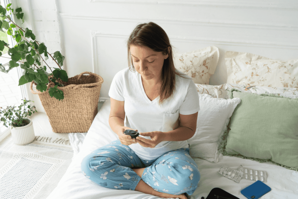 Woman sitting on bed in casual clothes checking her blood sugar level with a glucose meter in a bright bedroom.