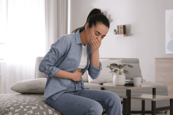 Young woman sitting on bed holding her stomach and covering her mouth, appearing nauseous.